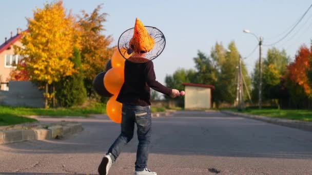 Halloween dzieci. uśmiechnięty chłopiec dynia twarz maska w kapeluszu czarownicy z pomarańczowymi i czarnymi balonami. Zabawne dziecko w strojach karnawałowych świętujące Halloween na świeżym powietrzu. Zwolniony ruch — Wideo stockowe