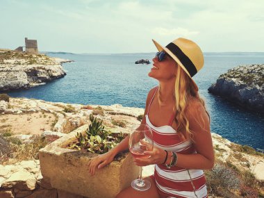 Young blonde woman in hat with drink enjoying the mediterranean feeling at seashore clipart