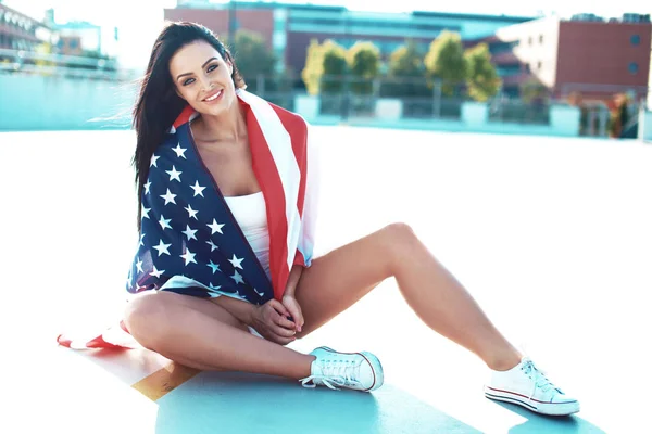 Happy young woman covered by USA flag sitting outdoors in sunlight