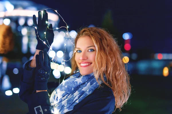 Happy young woman holding fairy lights at cold night in city, waiting for Christmas