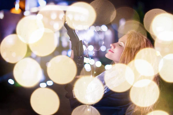 Young woman with fairy lights in cold city night imagine about Christmas, eyes closed