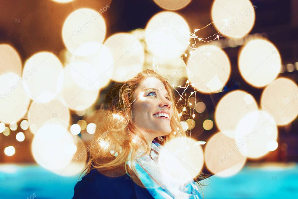 Happy young caucasian redhead woman amazed by LED fairy lights at city outdoor