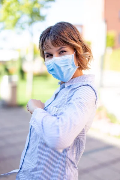 Young woman in mask greeting with elbow outdoors in park during pandemic