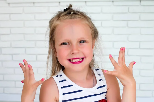 Little girl with her lips painted and shows her nails painted against a background with white bricks