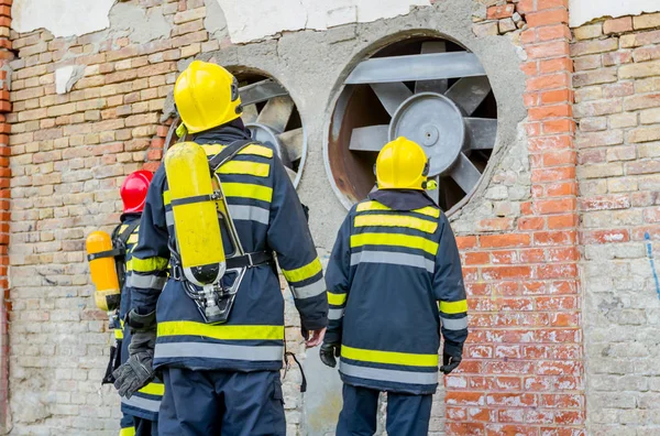 完全な安全ギヤと制服を着た消防士のチームの後ろから表示します 消防士がレンガの壁から来る空気トンネルに探しています — ストック写真