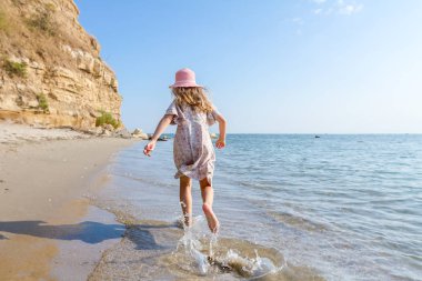 Genç kadın, çocuk şapka ve şirin yazlık elbise çalıştıran ve sığ su, deniz kıyısında.