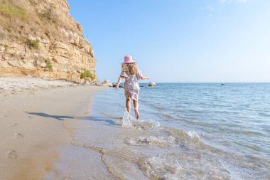 Genç kadın, çocuk şapka ve şirin yazlık elbise çalıştıran ve sığ su, deniz kıyısında.