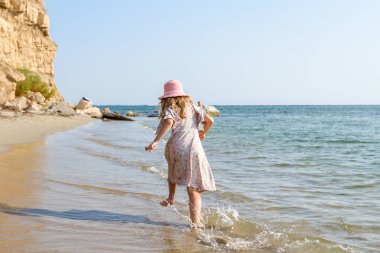 Genç kadın, çocuk şapka ve şirin yazlık elbise çalıştıran ve sığ su, deniz kıyısında.