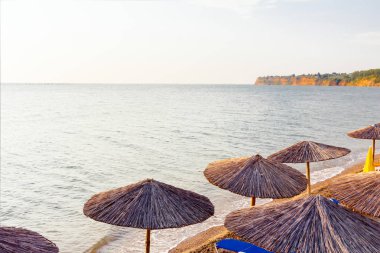 Sabahın erken saatlerinde güneş, şafak, halk plajında sazlı güneş gözlükleri, şemsiyeler ve deniz kıyısı boyunca iskemlelerle.