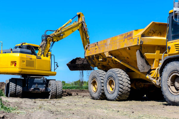 Big excavator is filling a dumper truck with soil at construction site, project in progress.