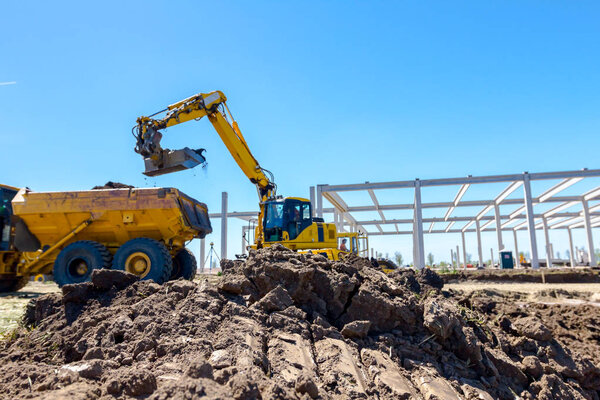 Big excavator is filling a dumper truck with soil at construction site, project in progress.