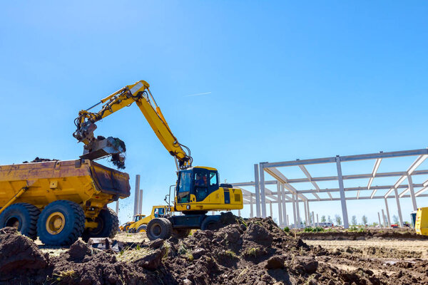 Big excavator is filling a dumper truck with soil at construction site, project in progress.