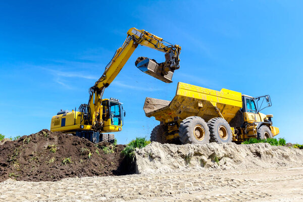 Big excavator is filling a dumper truck with soil at construction site, project in progress.