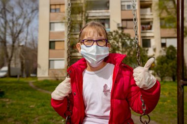 Baş parmakları havada, tıbbi maskeli ve eldivenli küçük bir kız COVID-19 virüsünden korunmak için tek başına sallanıyor.. 
