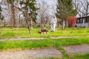 Koruyucu giysili bahçıvan bahçesinde çimen biçiyor. Çim biçme makinesi de meyve ağaçları arasında..
