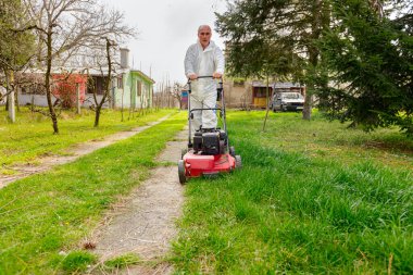 Koruyucu giysili bahçıvan bahçesinde çimen biçiyor. Çim biçme makinesi de meyve ağaçları arasında..