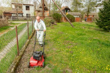 Koruyucu giysili bahçıvan bahçesinde çimen biçiyor. Çim biçme makinesi de meyve ağaçları arasında..