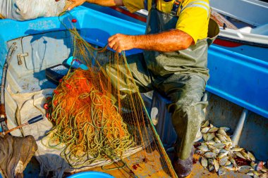 Kauçuk pantolonlu ve botlu balıkçı teknesinde oturuyor ve açık denizde balık tutmak için ağ topluyor..