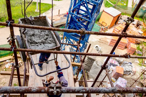 Workers are waiting crane to keep balance and direction to wheelbarrow, with mortar to placing on scaffold platform.