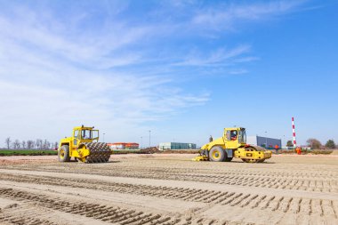İnşaat alanındaki kumu düzleştirirken, iki titreşimli buharlı silindirden oluşan ekip çalışması. Bir silindiri, plaka sıkıştırıcısını arkaya monte etti ve diğerinin zemine çarpmak için sivri uçlu çivileri var..