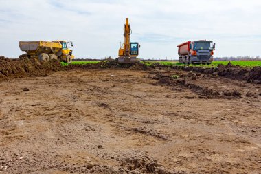 Büyük kazıcı iki çöp kamyonunu inşaat alanında toprakla dolduruyor..