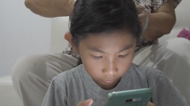 Menina Asiática Usando Smartphone Enquanto Sua Avó Fazendo Seu Cabelo — Vídeo de Stock