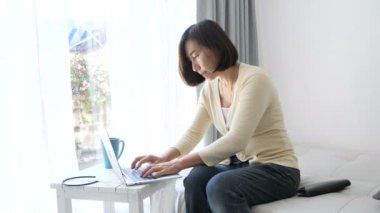 Asyalı kadın kanepede dizüstü evde pencere kullanarak rahatlatıcı.