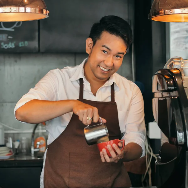 Happy Asian barista pouring milk into red coffee cup for latte art.