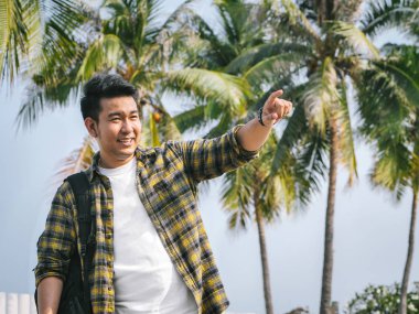 Hindistan cevizi ağacı arka plan ile deniz işaret eden sırt çantası ile Asya adam.