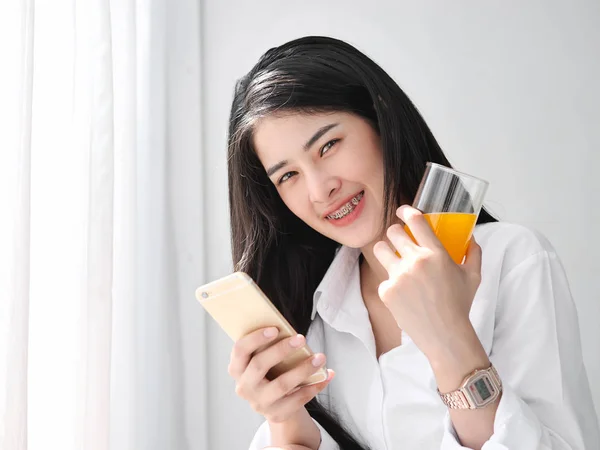 Mujer asiática feliz usando smartphone y sosteniendo un vaso de naranja —  Fotos de Stock