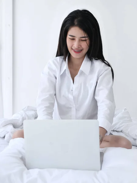 Mulher asiática feliz usando laptop na cama, relaxando em casa . — Fotografia de Stock