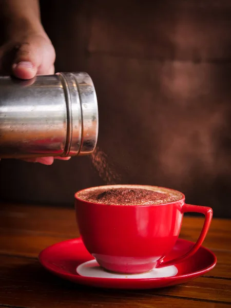 Ręka kropi czekolada w proszku na kubek kawy czerwony. — Zdjęcie stockowe