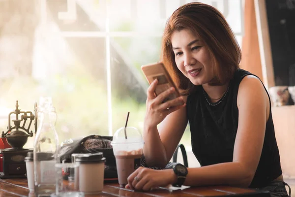 Mujer asiática bebiendo café y usando el teléfono inteligente en la cafetería . — Foto de Stock