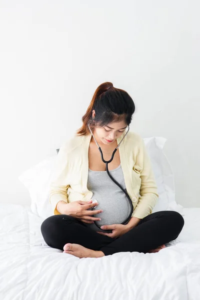 Беременная азиатка со стетоскопом слушает своего ребенка в постели — стоковое фото