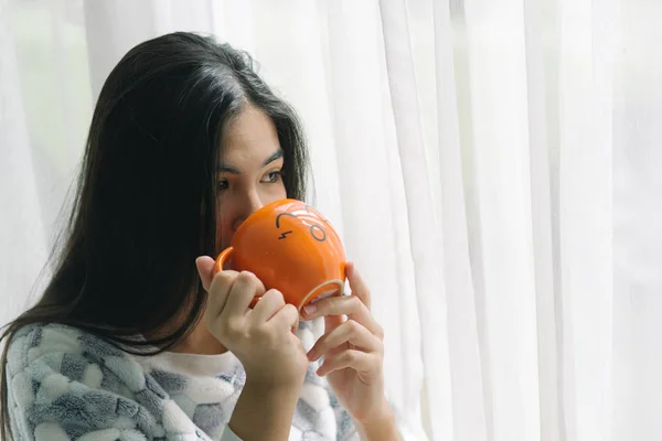 Asiatisk tonåring dricker lite dryck i orange kopp nära fönster, Li — Stockfoto