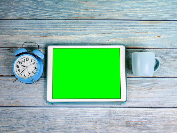 Ordenador tableta de pantalla verde, reloj despertador azul y taza de café sobre fondo de madera azul . — Foto de Stock