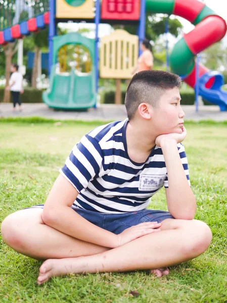 Nudne Azjatyckie preteen chłopak siedzi na zielonej trawie z placem zabaw — Zdjęcie stockowe