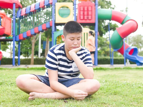 Langweilig asiatisch preteen junge sitting auf green grass mit spielplatz — Stockfoto