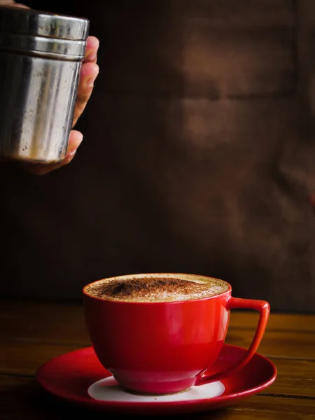 La mano espolvorea chocolate en polvo sobre la bebida en taza roja . —  Fotos de Stock