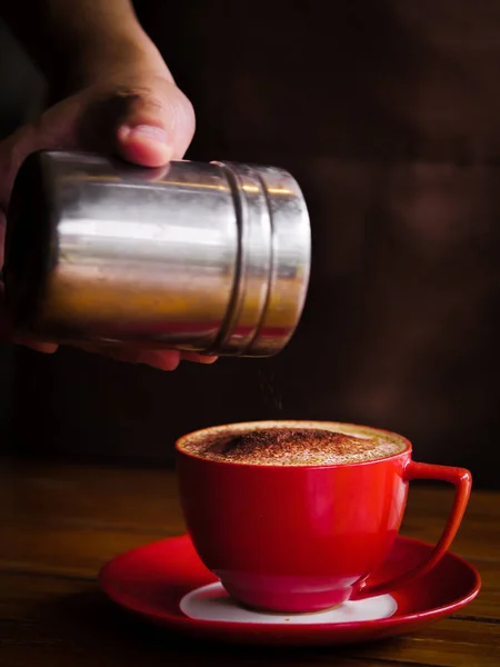 La mano espolvorea chocolate en polvo sobre la bebida en taza roja . —  Fotos de Stock