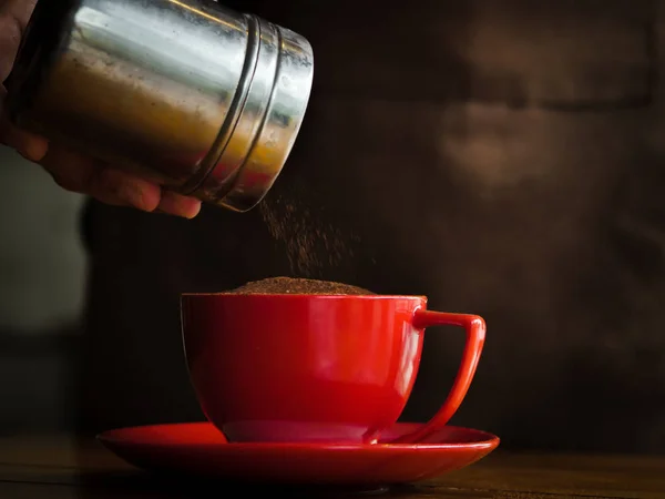 La mano espolvorea chocolate en polvo sobre la bebida en taza roja . —  Fotos de Stock