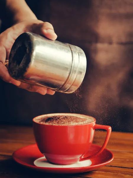 La mano espolvorea chocolate en polvo sobre la bebida en taza roja . —  Fotos de Stock