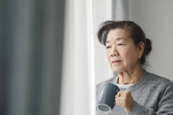 Asian senior woman drinking hot tea near window outdoor, lonely — Stock Photo, Image