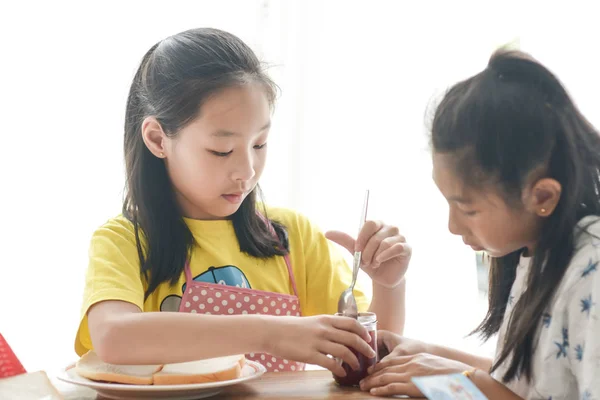Asian sister spreading strawberry jam on bread for her younger s
