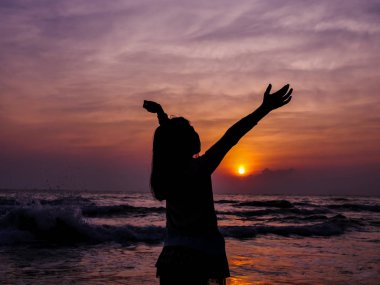 Plaj arka planda güneş doğarken el kaldıran kız.