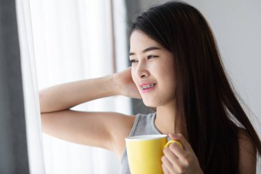 Asyalı kadın doğal ışık ile pencere yakınındaki sarı kupa holding.