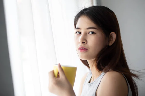 Mujer asiática sosteniendo taza amarilla cerca de ventana con luz natural . —  Fotos de Stock