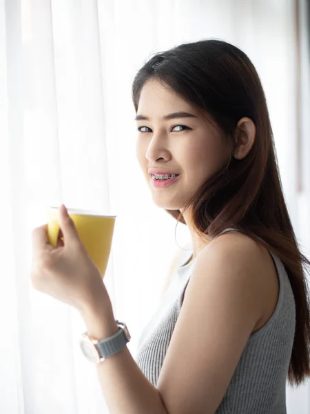 Mujer asiática sosteniendo taza amarilla cerca de ventana con luz natural . —  Fotos de Stock