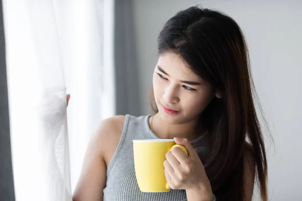 Mujer asiática sosteniendo taza amarilla cerca de ventana con luz natural . —  Fotos de Stock