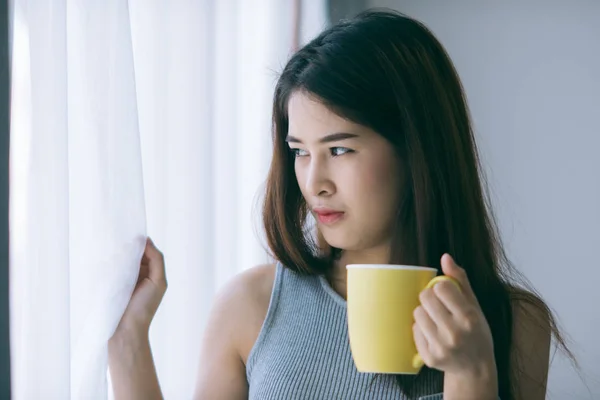 Mujer asiática sosteniendo taza amarilla cerca de ventana con luz natural . —  Fotos de Stock